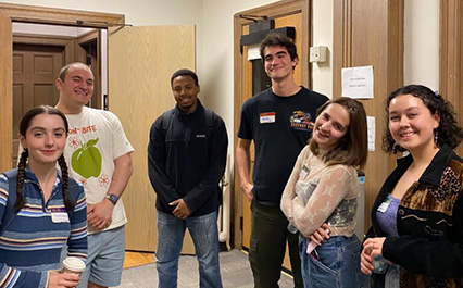 Six students smile at a campus event, wearing name tags and casual clothes inside a classroom hallway.