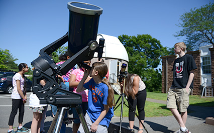 Children and teens use telescopes outside during a sunny public astronomy outreach event.