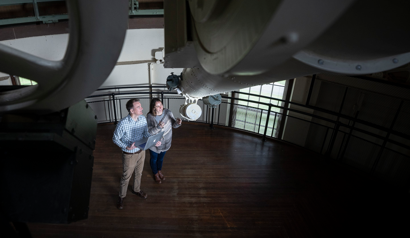 Two people, a man and a woman, stand inside an observatory, looking up at a large telescope mounted above them. The man holds a laptop, and both appear engaged and focused. 