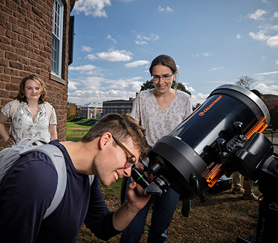 A student peers into a telescope outdoors while two others watch; middle
