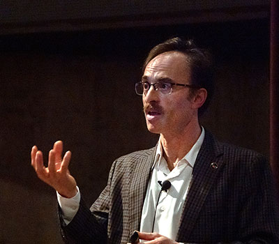 Dr. Sheperd S. Doeleman—Astrophysicist at the Center for Astrophysics | Harvard & Smithsonian and Director of the Event Horizon Telescope (EHT), wearing glasses and a checked blazer gestures during a lecture in a dimly lit room.