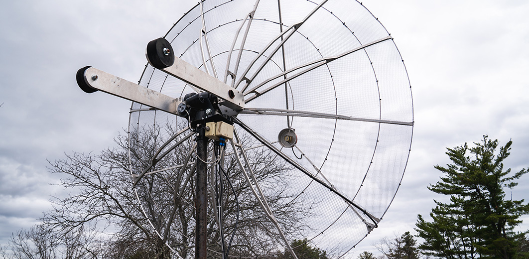 A large mesh radio dish built by students points skyward on a cloudy day, surrounded by trees.