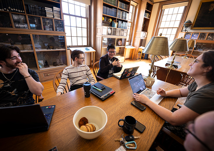 Students gather around a table with laptops and notebooks in a bright, book-filled library room.