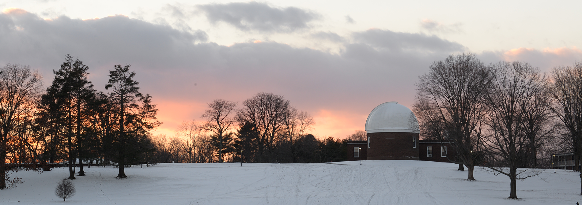 A snowy winter scene of the Wesleyan observatory surrounded by trees at dusk, under a cloudy purple-orange sky.