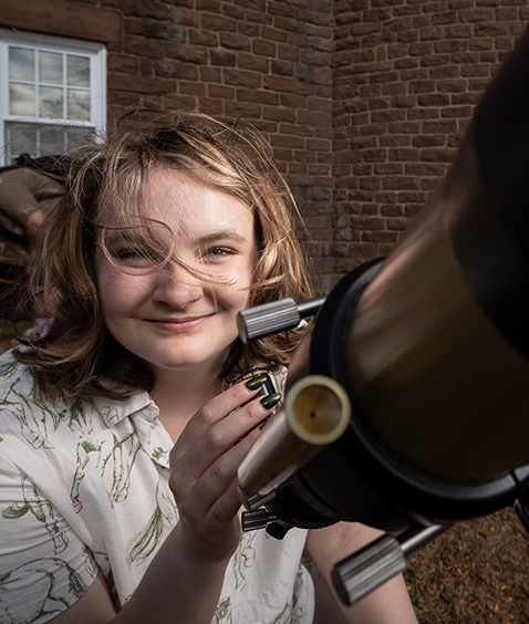 Portrait of a student with windblown hair smiling as she looks through a telescope outdoors during daylight.