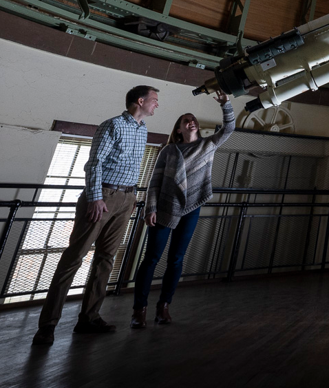 Two students inside an observatory, a man and a woman, looking up at a large telescope suspended overhead, the room dimly lit.