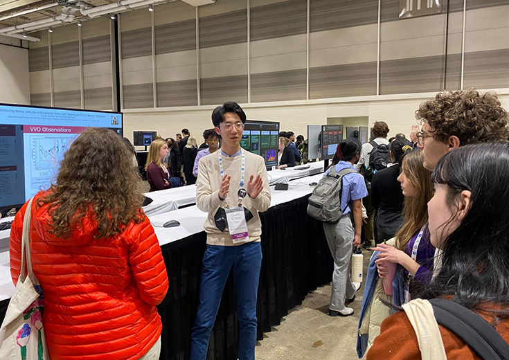 Student presents astronomy research at a conference, speaking to a group in front of digital posters.