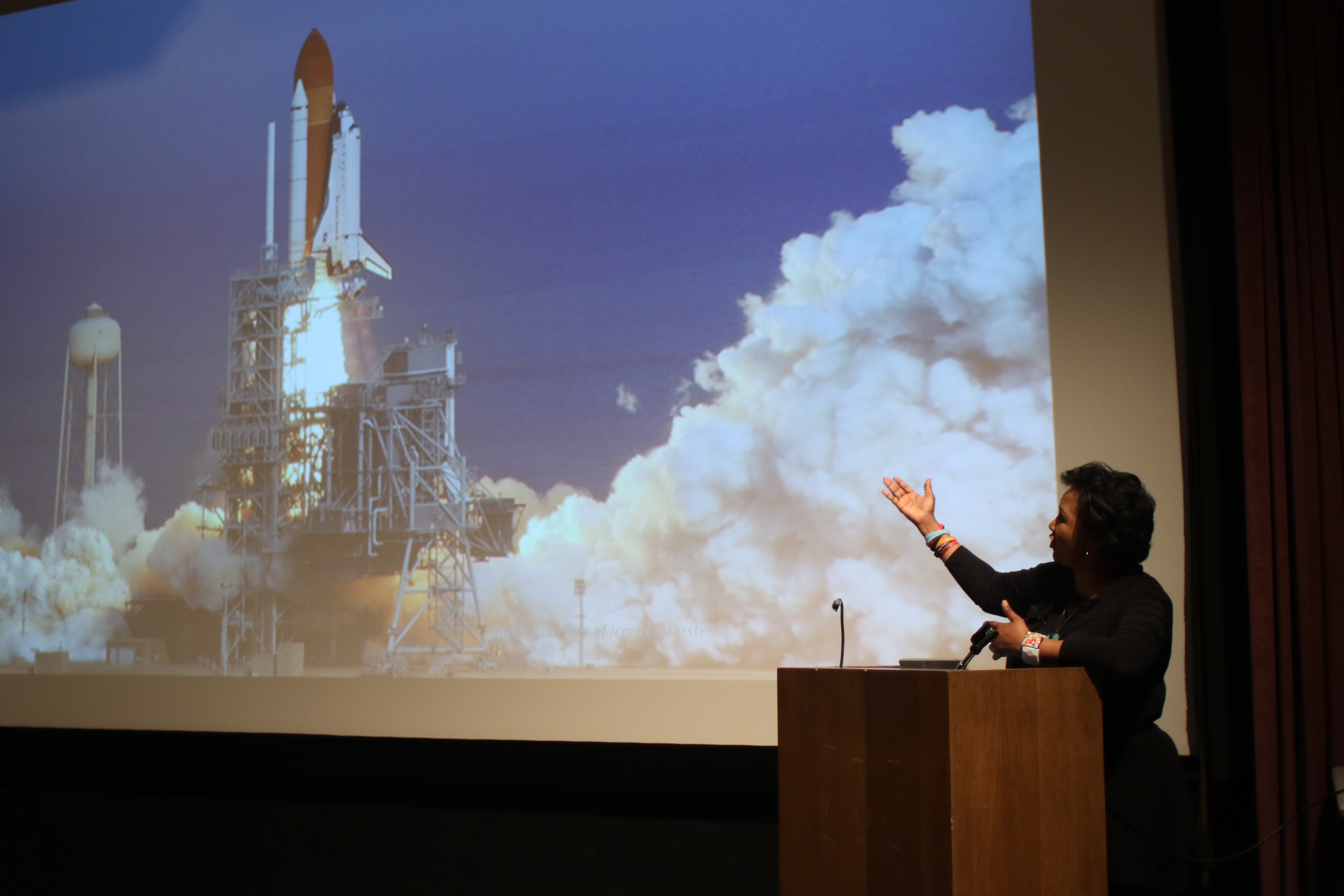 speaker at a podium pointing to a projected image of a rocket launch